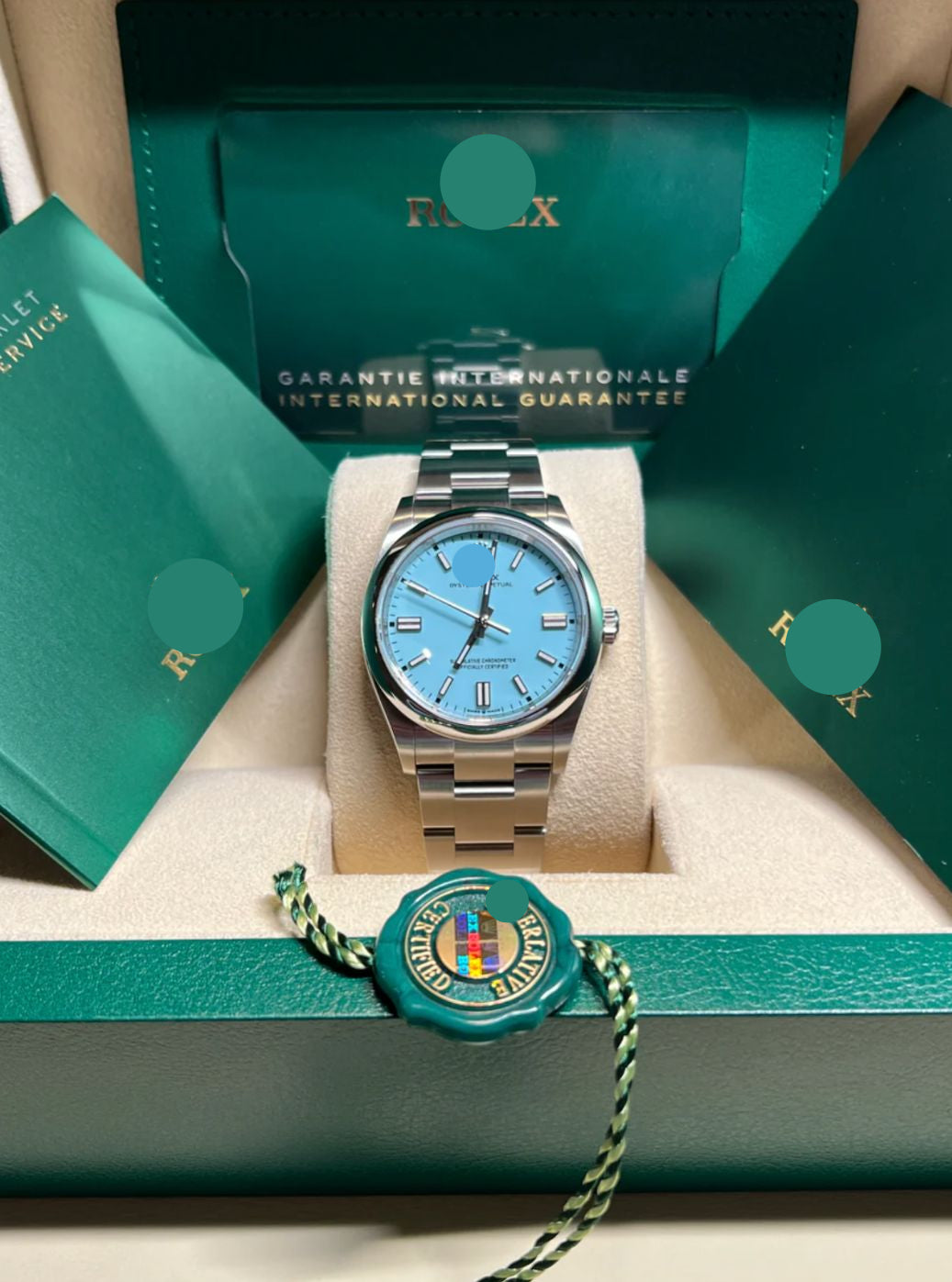 Rolex watch with blue face and silver case on a green cushion, surrounded by Rolex packaging.